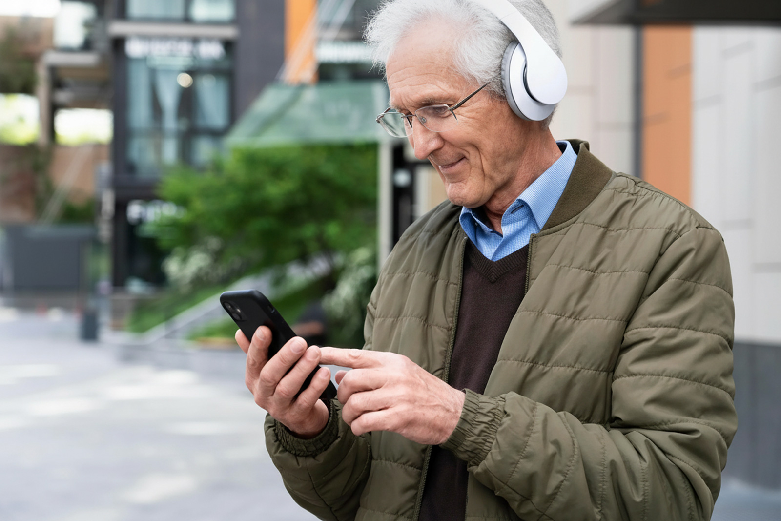 An older man standing outdoors on a city sidewalk wearing headphones, smiling and using a smartphone.